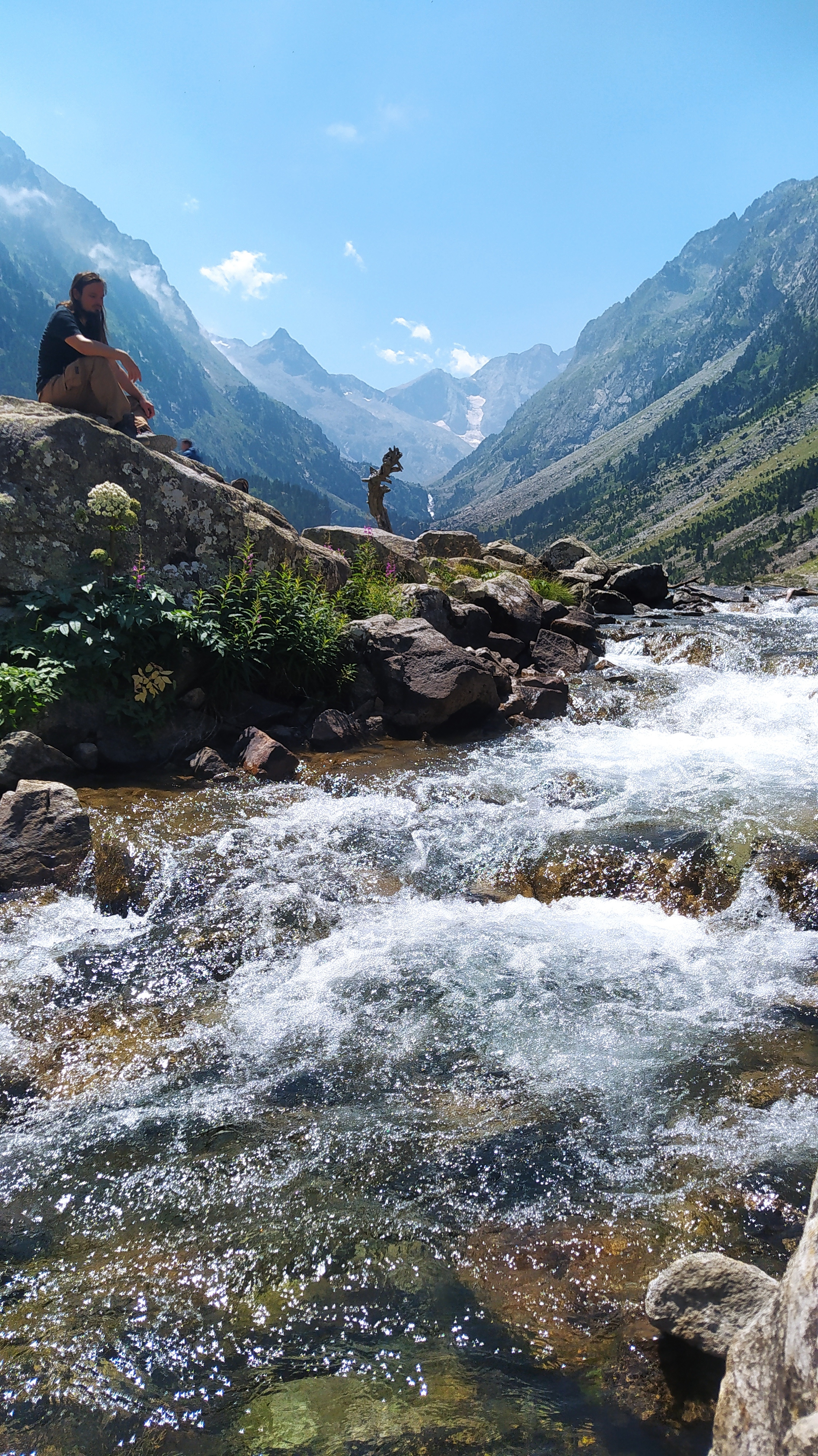 Photo d'une rivière surplombée par une magnifique montagne d'été. Noé apparaît perché sur un rocher, pensif.